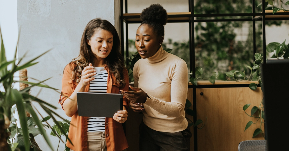 Two women discuss something on a tablet in a modern office filled with plants